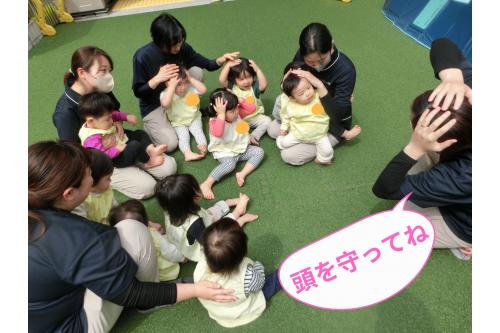 揺れがおさまった後は、<br><span style='color:#ff7f00;'>安全を確保</span>した園庭に避難しました。<br><br><span style='color:#7f00ff;'>園庭でも頭を守って</span>靴や安全帽を待ちます。<br><br>子ども達もとっても上手に<br>頭を守ることができました。<br>